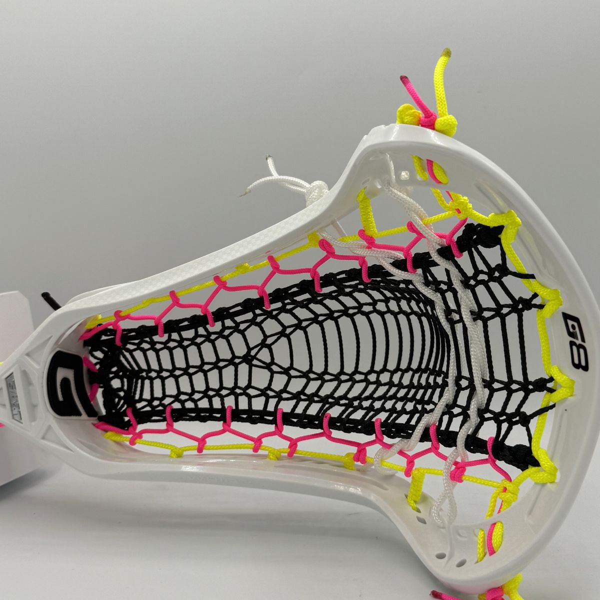Close-up of a lacrosse head with white frame, black and pink strings, and yellow accents on a gray background.